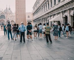 A group of tourists enjoys the benefits of escorted tours in a historic square, surrounded by ornate buildings. In the background is a tall brick tower, likely part of a cathedral. The scene is bright, suggesting daytime, and people wear casual, summer clothing.