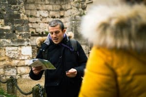 A man in a black coat with a fur lined hood is reading a map outside. He is standing near a stone wall, and a person in a yellow jacket is in the foreground, slightly blurred. It appears to be a cool day. Senlis, France