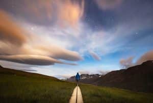 A person in a blue jacket stands on a wooden path, gazing at a star filled sky with dramatic, swirling clouds over Akureyri. The landscape is a grassy field with the majestic mountains of the North in the background.