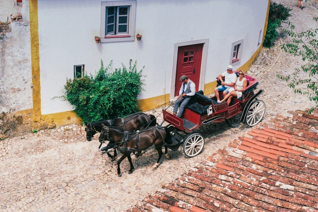A red horse drawn carriage with a driver and three passengers travels on a cobblestone street alongside a white building with red door and windows. Lush greenery and terracotta roof tiles are visible in the scene.