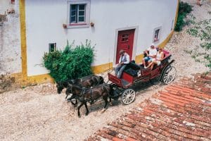 A red horse drawn carriage with a driver and three passengers travels on a cobblestone street alongside a white building with red door and windows. Lush greenery and terracotta roof tiles are visible in the scene.