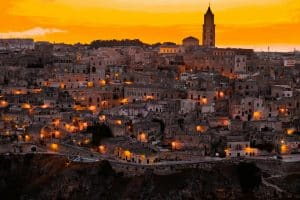 A stunning view of Matera at sunset, with the old city's stone buildings illuminated by warm, golden lights. A church tower rises prominently against the vibrant orange sky, creating a picturesque scene of this historic Italian city.