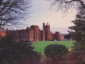 A large, historic brick building with tall towers and detailed architecture stands on a lush green lawn. The scene is framed by trees and shrubs, with soft, overcast lighting in the sky.