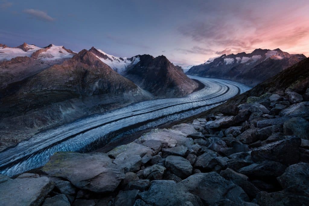 A winding glacier stretches through a rocky mountain range at dusk. The sky transitions from blue to pink, casting a serene glow over the landscape. Rugged stones are scattered in the foreground, enhancing the natural beauty.