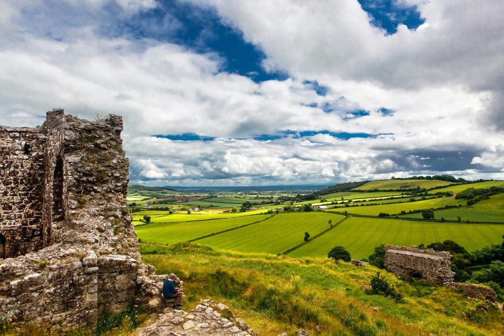 Ruins of an ancient stone structure sit atop a lush, green hill with grassy fields extending into the distance. A person in blue relaxes near the ruins. Puffy white clouds float in a vibrant blue sky, completing the picturesque landscape.