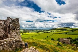 Ruins of an ancient stone structure sit atop a lush, green hill with grassy fields extending into the distance. A person in blue relaxes near the ruins. Puffy white clouds float in a vibrant blue sky, completing the picturesque landscape.