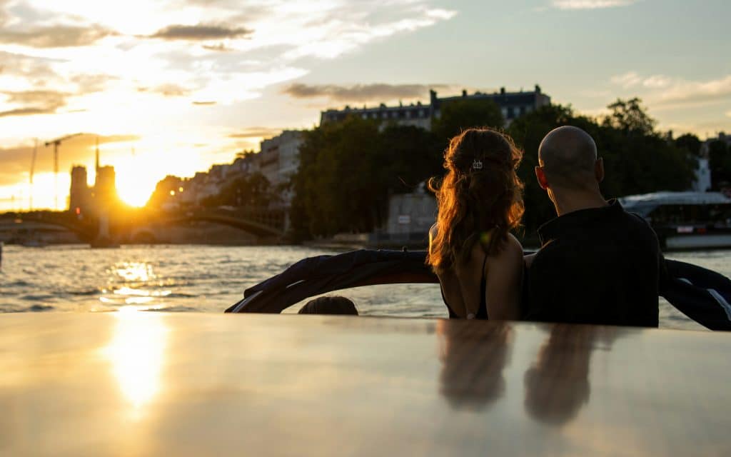 A couple sits at the back of a boat, watching the sunset over a river. The sun casts a warm glow on the water and silhouettes the couple. Buildings and trees line the riverbanks under a partly cloudy sky. Paris, France