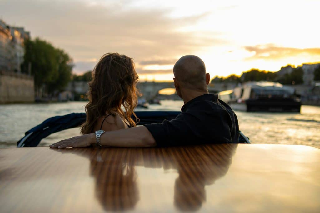 A couple sits on a boat, overlooking a sunset on a river. The man's arm is around the woman as they enjoy a serene view of the water, surrounded by other boats and city buildings.