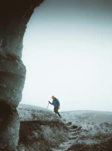 A person wearing an orange helmet and blue jacket stands on an icy terrain, holding a walking stick. The sky is overcast and a large ice formation is partially visible on the left.