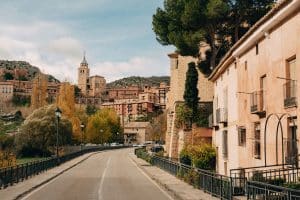 Scenic view of a winding road leading to a historic town with rustic buildings and a prominent church tower. Trees with autumn foliage line the road, and the background features hills under a partly cloudy sky. Albarracín, Spain