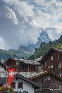 Swiss village scene with traditional wooden houses, one displaying a Swiss flag. The background features a dramatic view of the Matterhorn partially obscured by clouds. Red flowers decorate the balconies, adding color to the rustic setting.