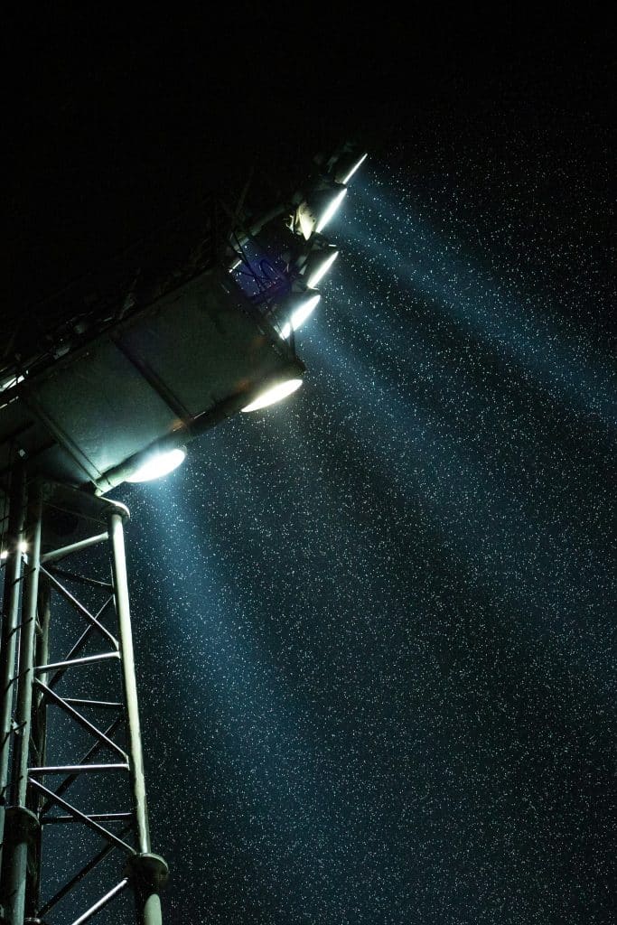 A tall metal structure with bright spotlights illuminates the falling rain against a dark night sky, creating a dramatic scene reminiscent of the 2025 NFL Dublin Game, where beams of light cut through the raindrops like players slicing through defense.