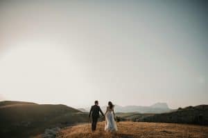 A couple in formal attire walks hand in hand on a grassy hillside at sunset, reminiscent of a private group tour admiring nature’s beauty. The expansive landscape of rolling hills and distant mountain range under a clear sky creates a romantic and serene atmosphere.