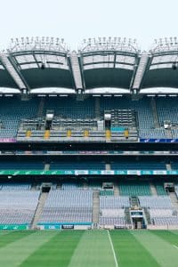 View of an empty sports stadium, featuring multiple tiers of seating. The field is well maintained with a clear white line in the center. Stadium lighting and a partial roof structure are visible at the top. Croke Park, Dublin, Ireland