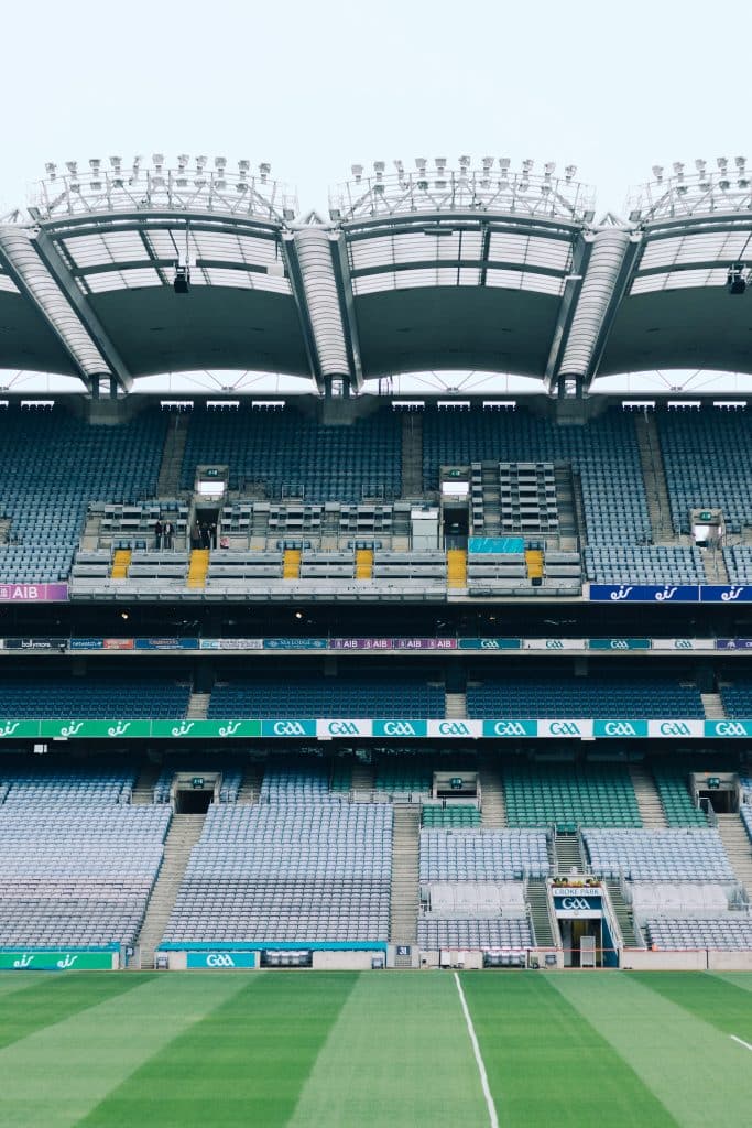 View of an empty sports stadium, featuring multiple tiers of seating. The field is well maintained with a clear white line in the center. Stadium lighting and a partial roof structure are visible at the top. Croke Park, Dublin, Ireland