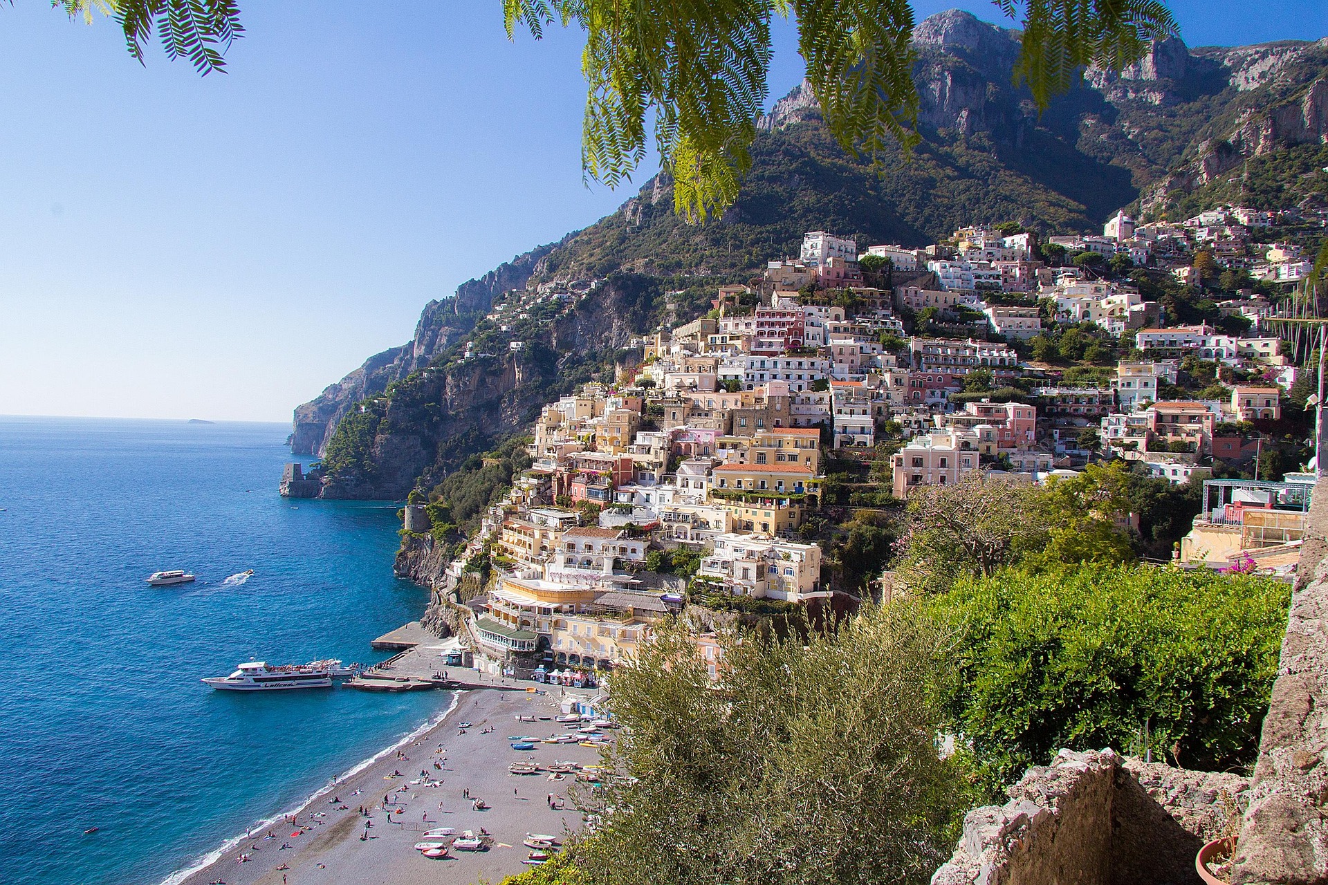 Colorful buildings cascade down a steep hillside toward a beach with umbrellas and boats on bright blue water in Positano, Italy, under a clear sky, with lush greenery—an iconic view perfect for Amalfi Coast custom itineraries.