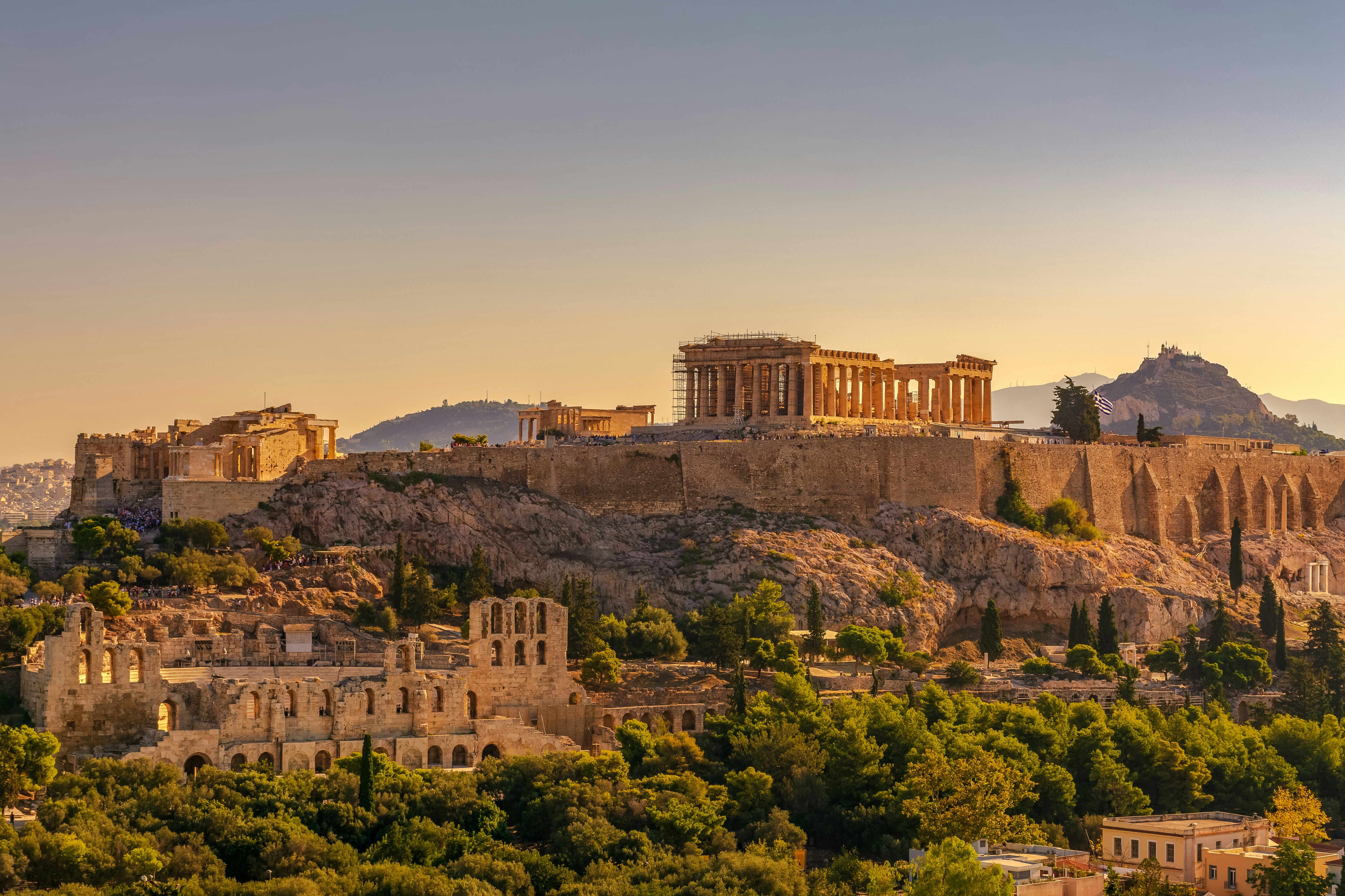 The Acropolis of Athens at sunset, with the Parthenon and ancient ruins on a rocky hilltop, offers a romantic view perfect for those inspired by England & Ireland Honeymoon Packages, surrounded by greenery and cityscape under a clear sky.
