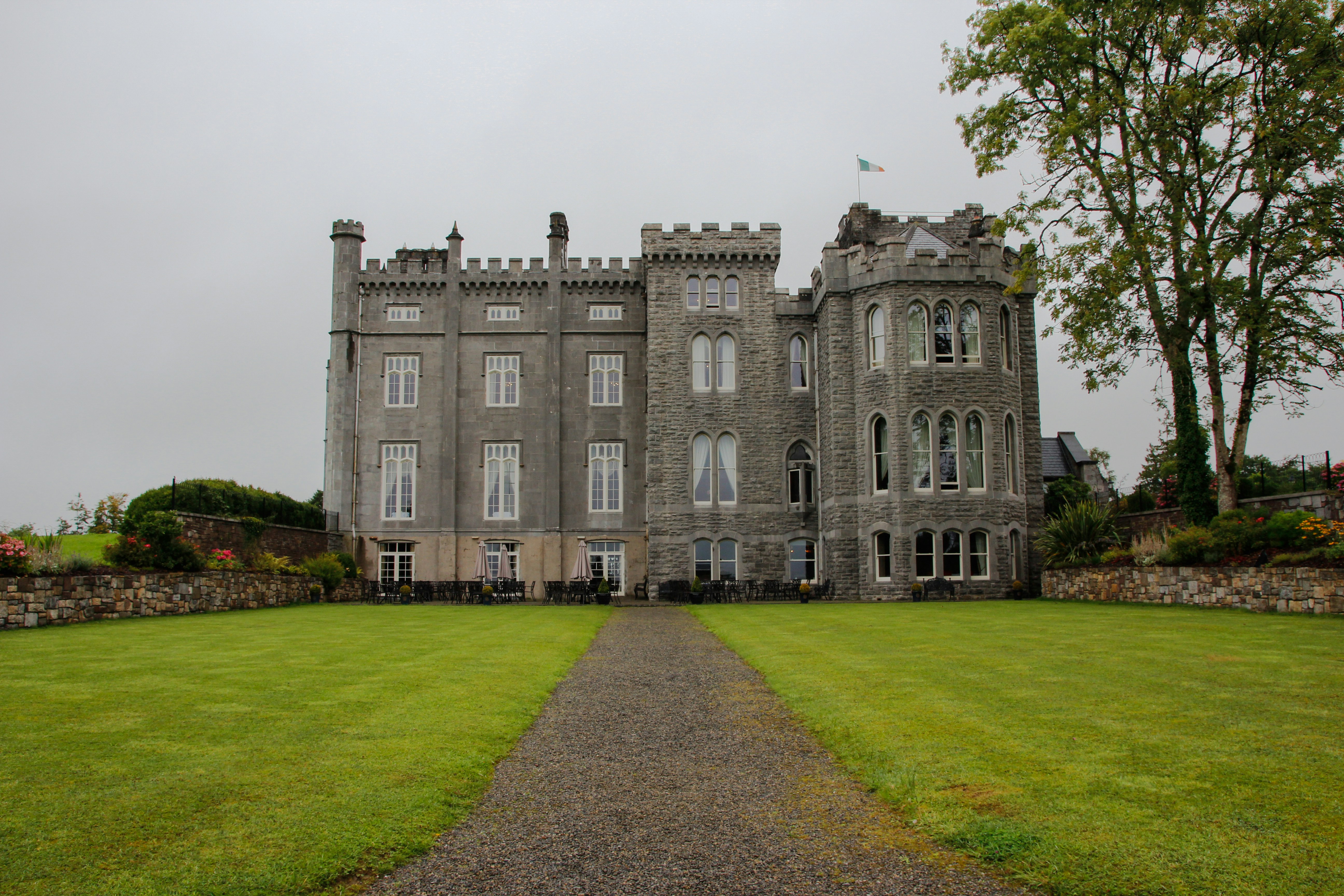 A large, gray stone castle with tall windows, battlements, and a central tower stands at the end of a gravel path, surrounded by neatly trimmed lawns and a stone wall on a cloudy day.