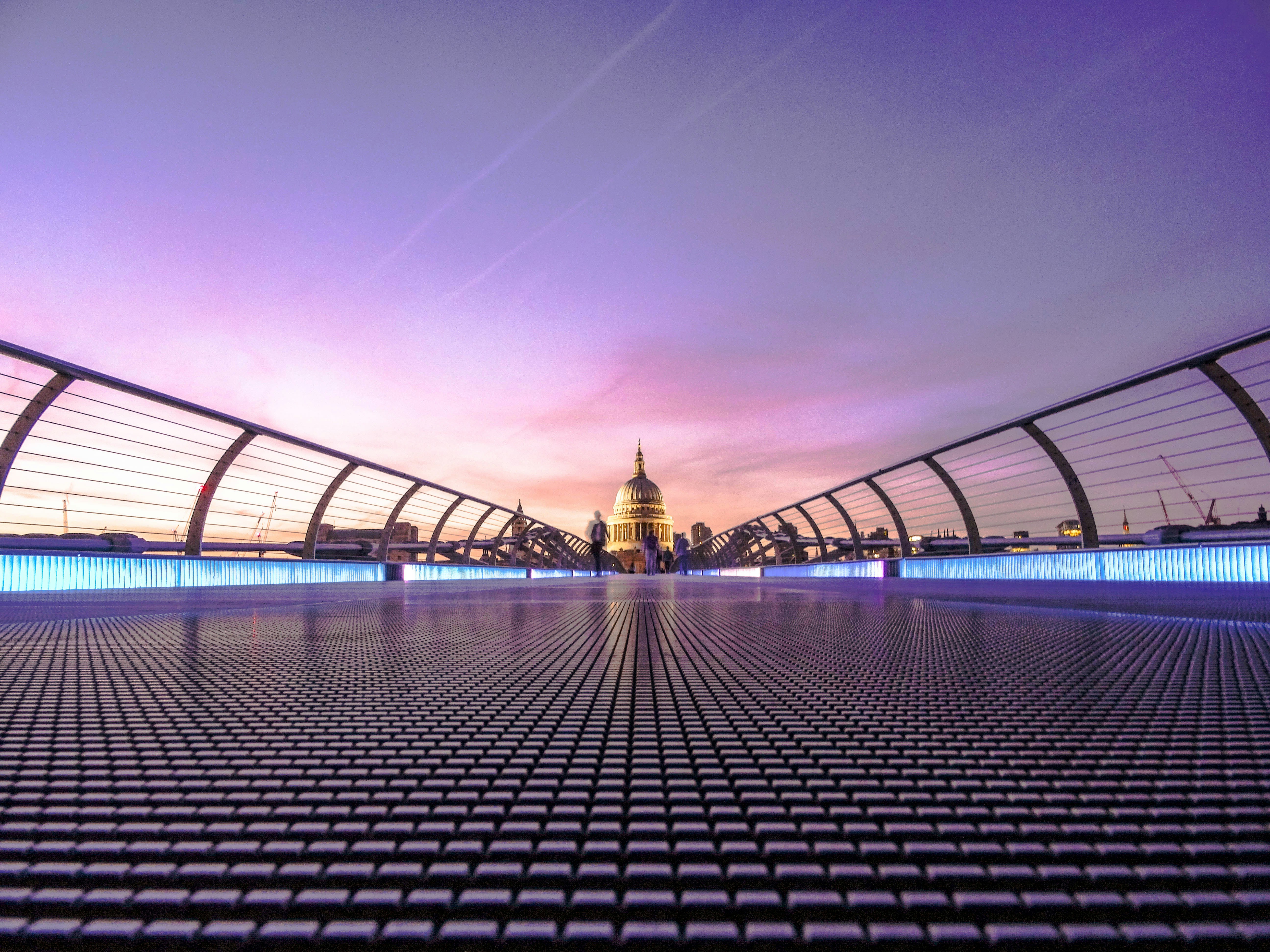A low angle view of the Millennium Bridge in London at sunset, with the dome of St. Paul’s Cathedral centered in the background under a purple and pink sky.
