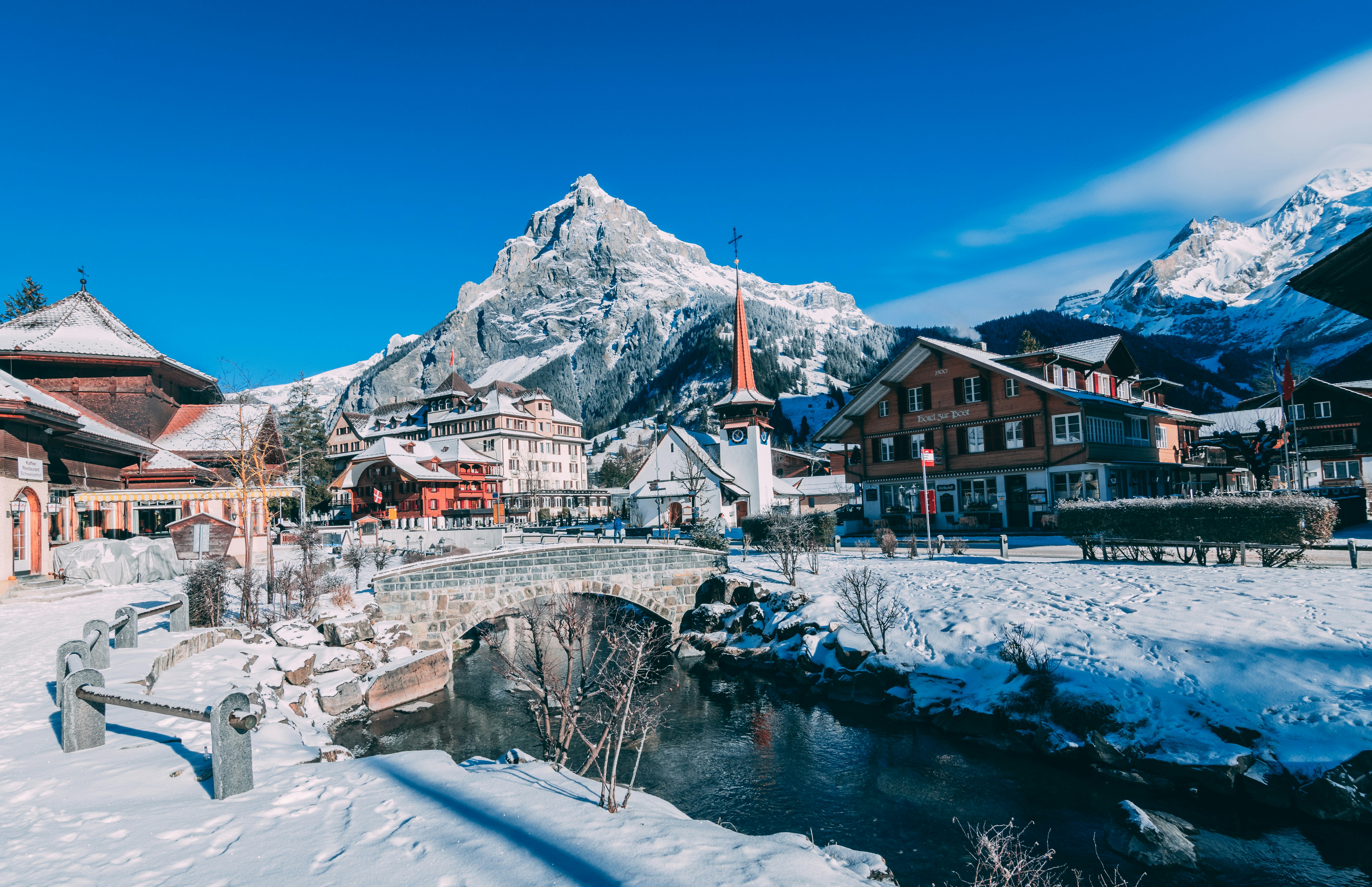 A picturesque snowy village with Alpine style houses, a church steeple, stone bridge, and stream, set against snow covered mountains—perfect for inspiring your Italy and Greece Honeymoon Itinerary under a clear blue sky.