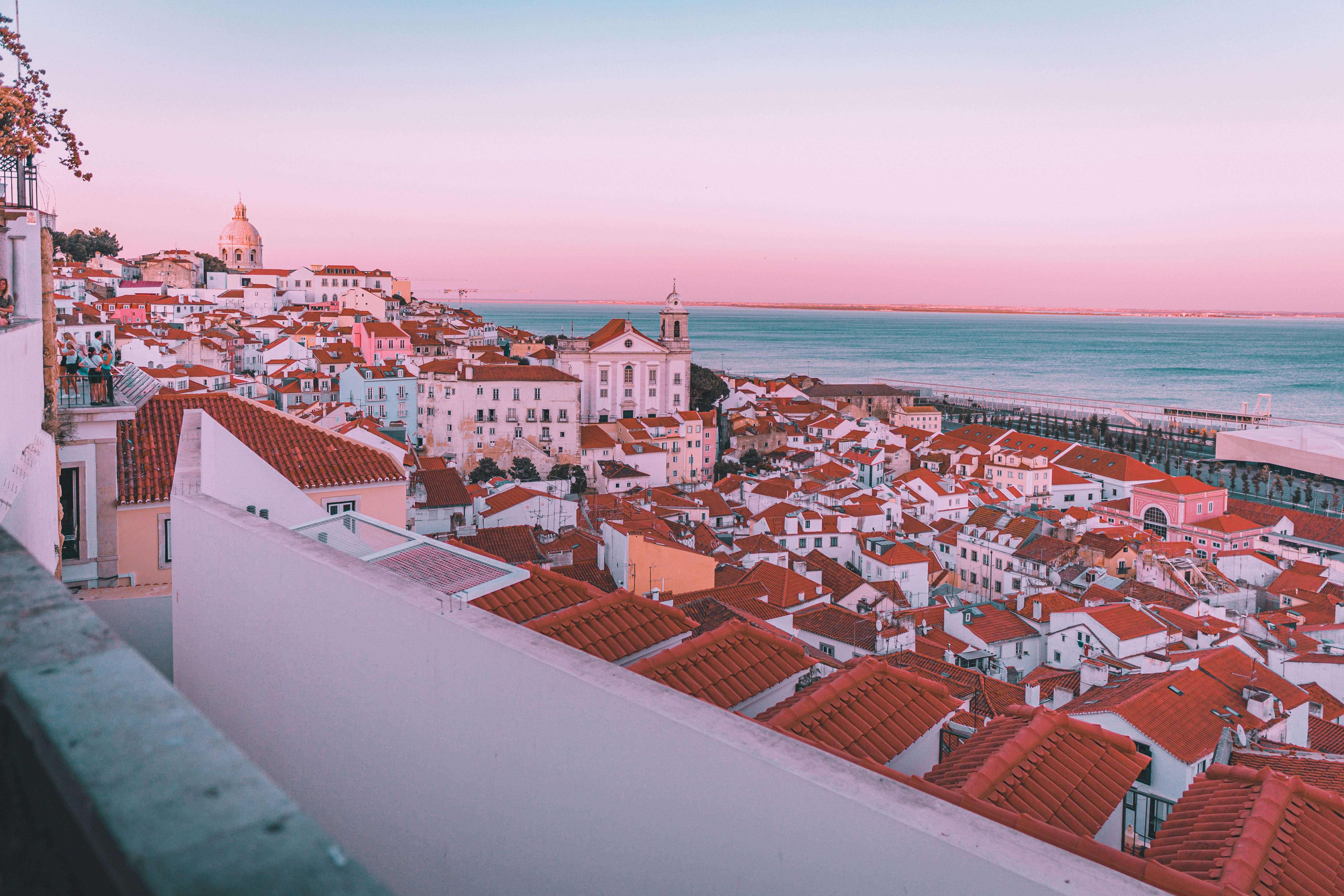 A panoramic view of Lisbon’s Alfama district at sunset, showing red tiled rooftops, pastel colored buildings, a church, and the Tagus River under a pink and blue sky.