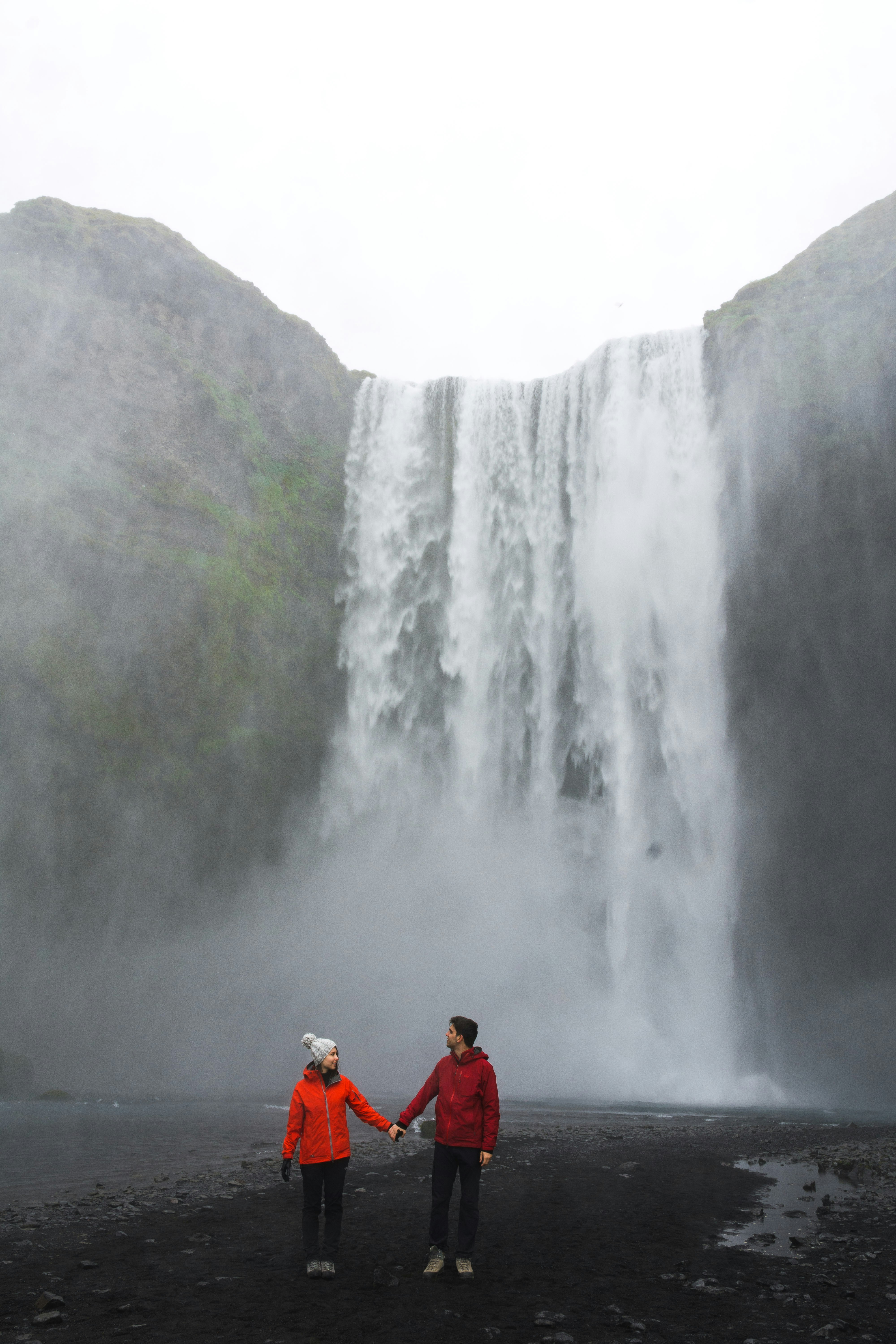 Two people in red jackets hold hands before a misty waterfall, rocky cliffs, and cloudy sky—an enchanting scene inspired by a Guide to Honeymoon in France.