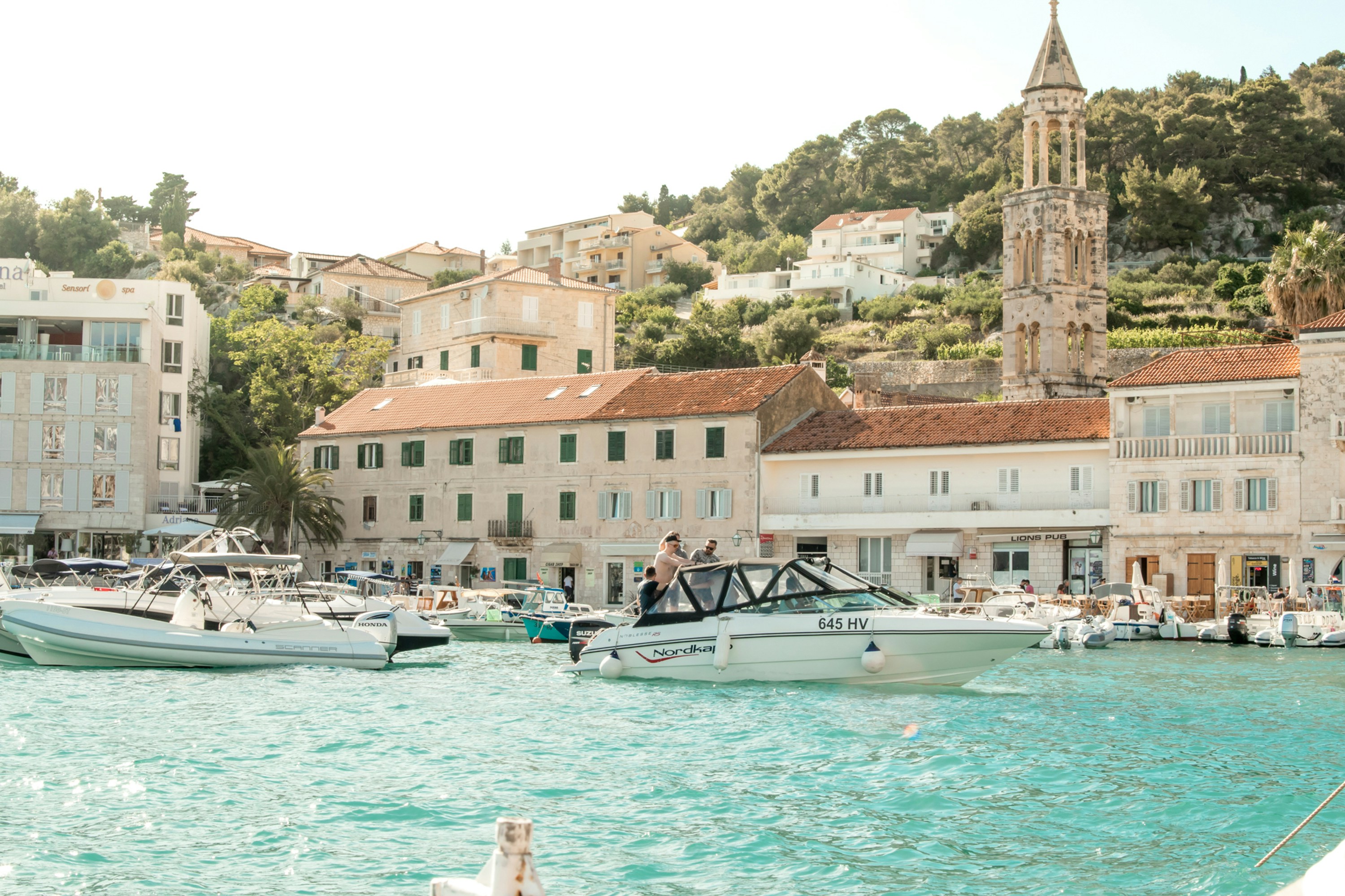 A small motorboat with people onboard floats on turquoise water near a marina, surrounded by other boats—an idyllic scene often featured in Mediterranean Honeymoon Packages. Historic buildings, a church tower, and green hills complete the background. Hvar, Croatia