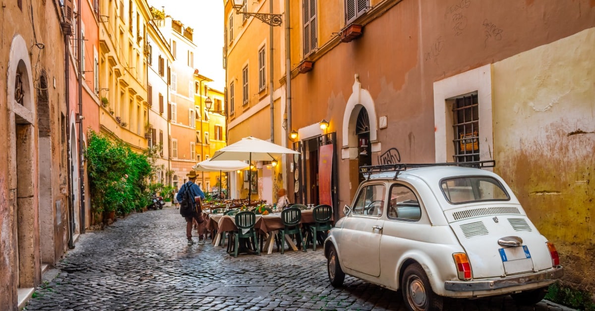 A tight alleyway in Italy with local cafe tables and umbrellas off to the side. There is a car parked behind the dining area.