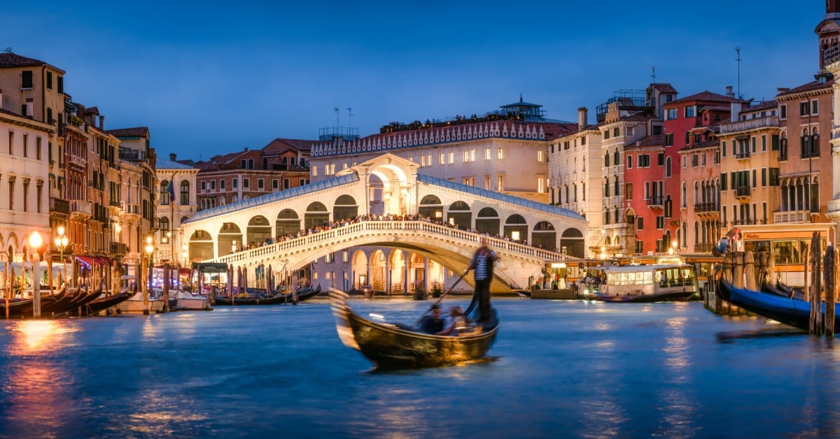A night time view of a bridge in Italy with a romantic gondola ride happening in the middle of the river.